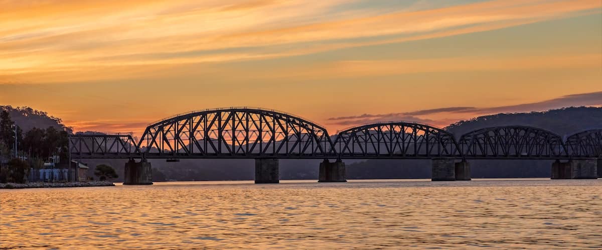 A steel bridge spanning a river at sunset with a mountainous backdrop.