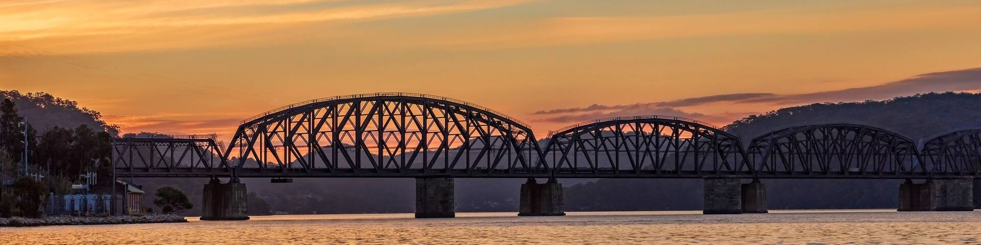 A steel bridge spanning a river at sunset with a mountainous backdrop.