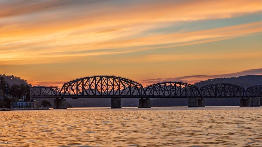 A steel bridge spanning a river at sunset with a mountainous backdrop.