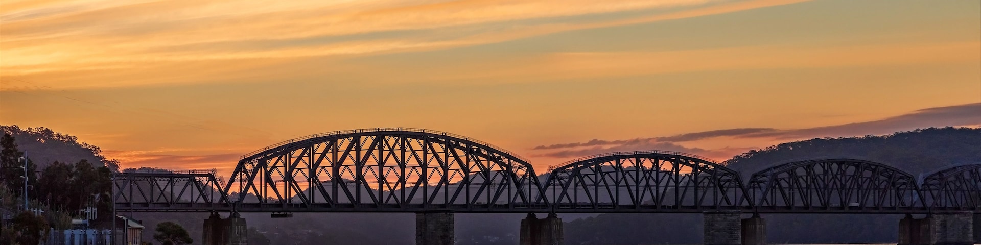Ponte de aço atravessando um rio ao pôr do sol, com montanhas ao fundo.