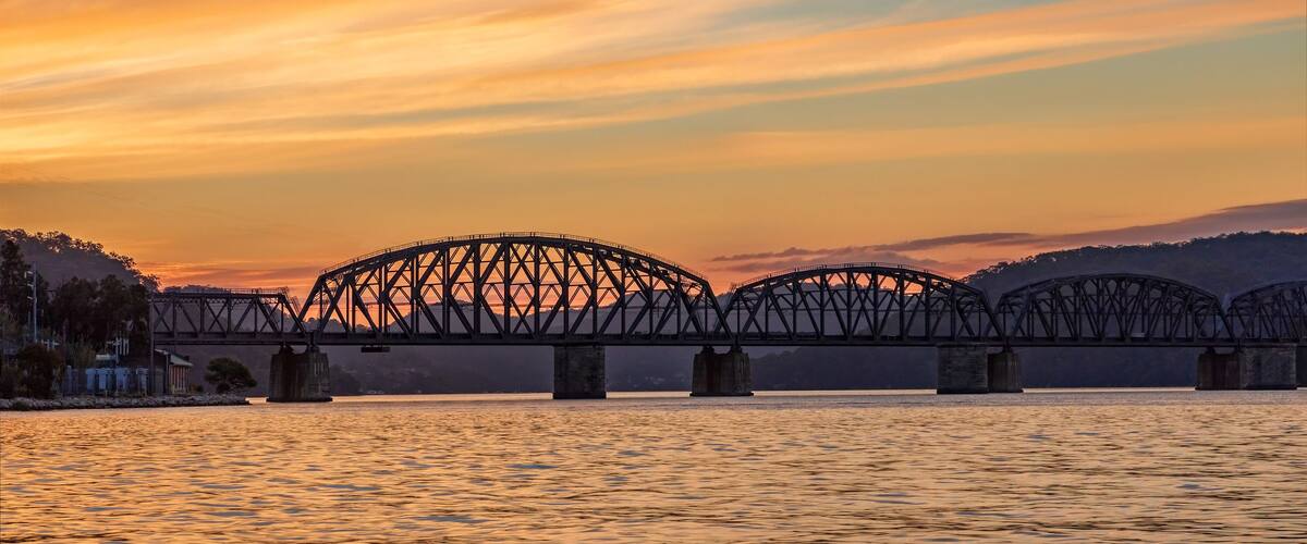 A steel bridge spanning a river at sunset with a mountainous backdrop.