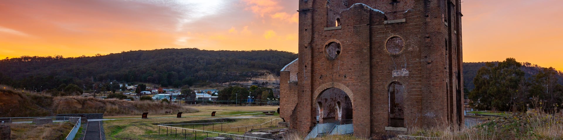 A historic brick tower with arched windows and a staircase leading up to it, surrounded by grassy fields and a sunset sky.