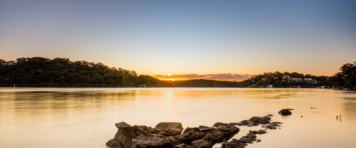 A calm lake at sunset with a rocky shoreline in the foreground and a forested hill in the background.