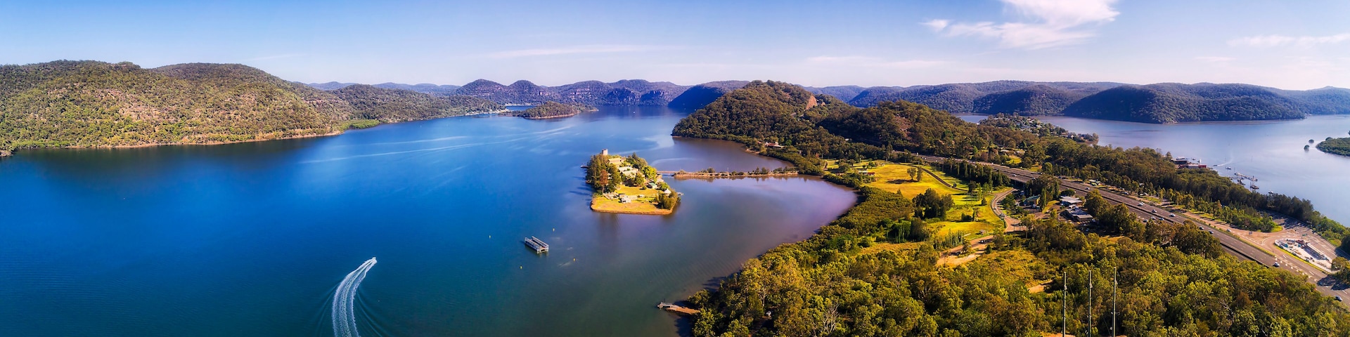 Um lago extenso cercado por colinas, uma pequena ilha e uma estrada com veículos.