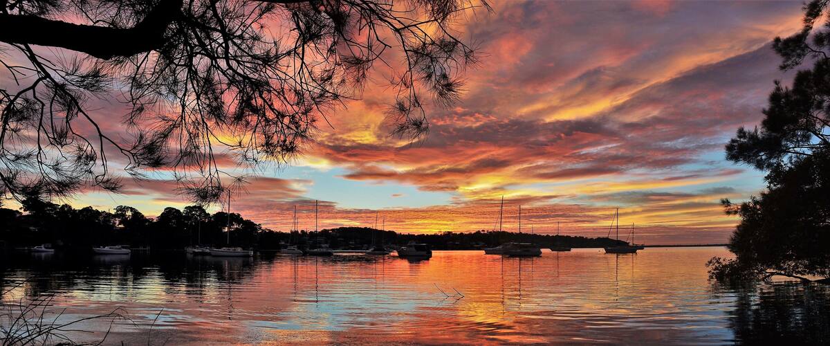 A sunset over a calm lake with boats, trees, and a distant shoreline.
