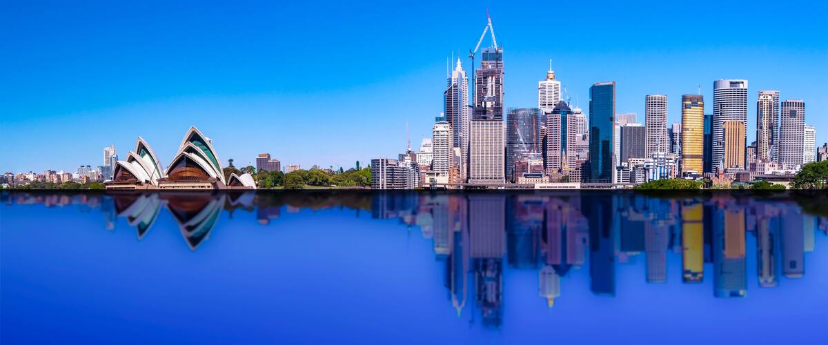 A city skyline with modern skyscrapers and the Sydney Opera House reflected in the water.