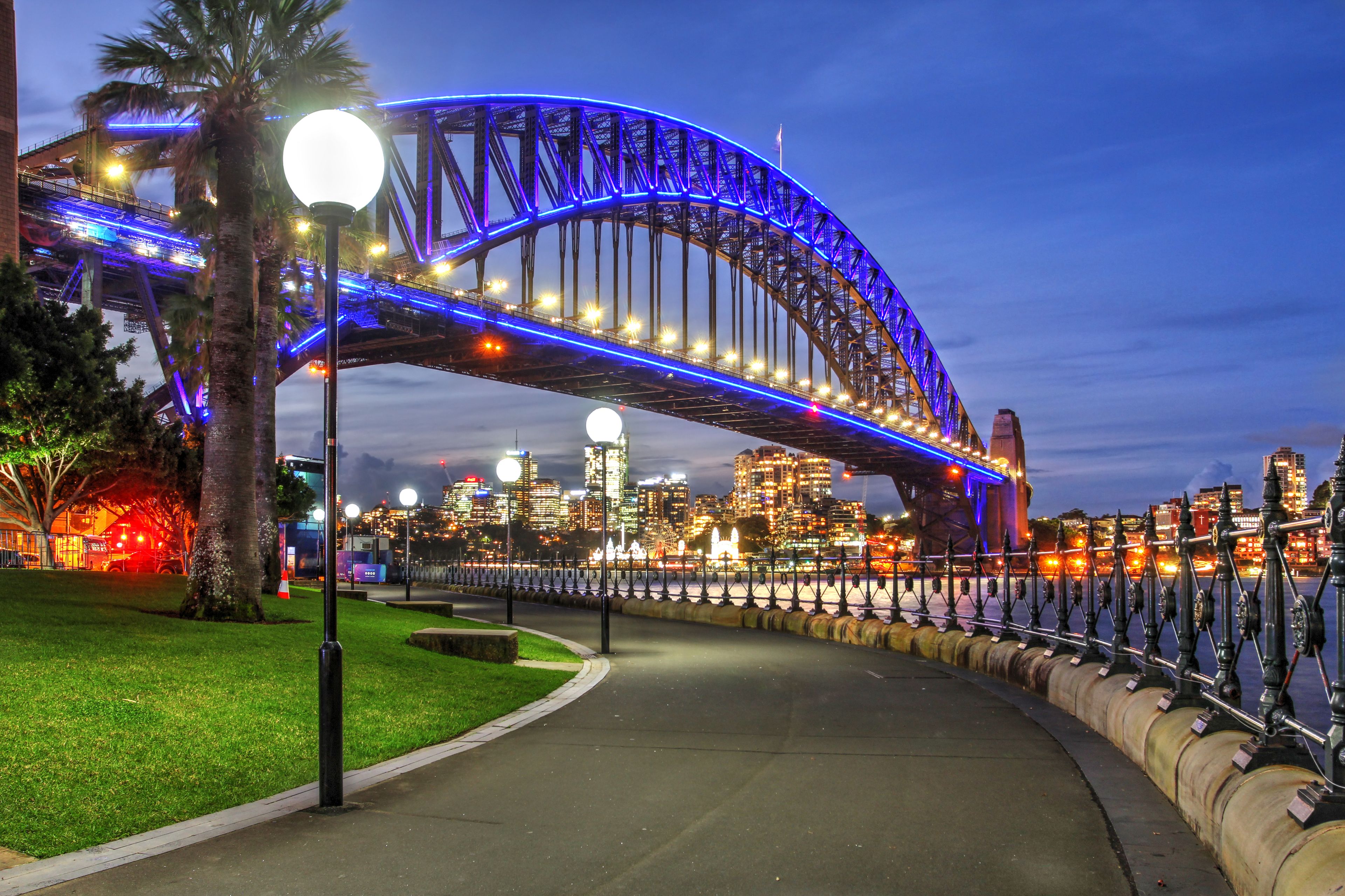 A brightly lit bridge with blue lights, a city skyline in the background, and a pathway with street lamps.