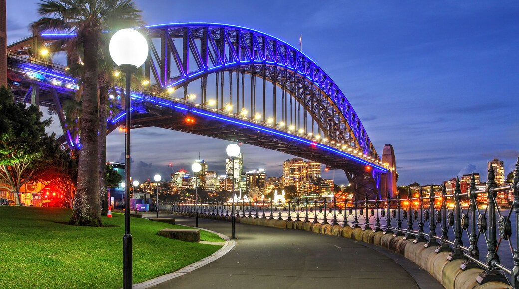 A brightly lit bridge with blue lights, a city skyline in the background, and a pathway with street lamps.