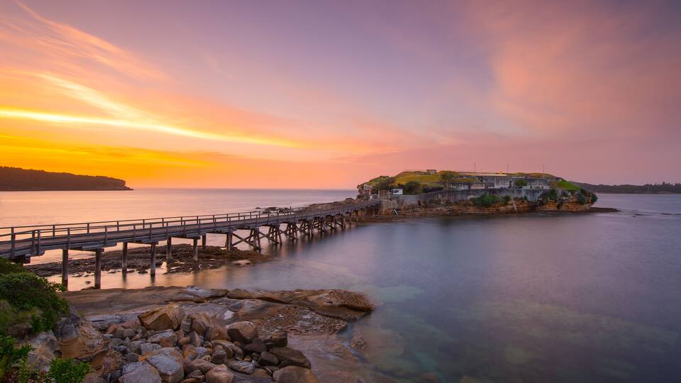 A coastal sunset with a pier leading to a fortified island.