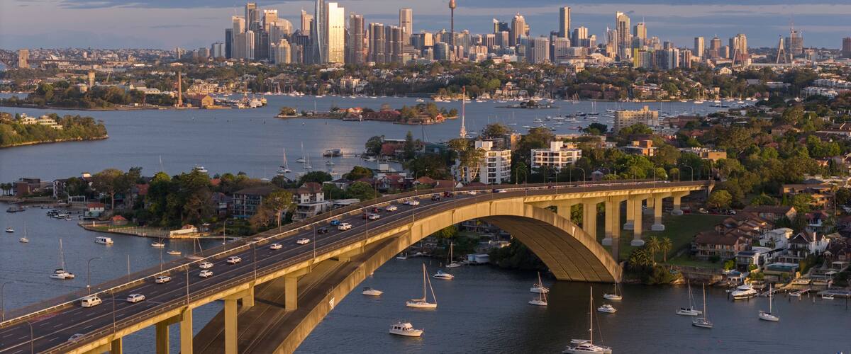 A bridge over a river with boats, a city skyline in the background, and a clear sky.