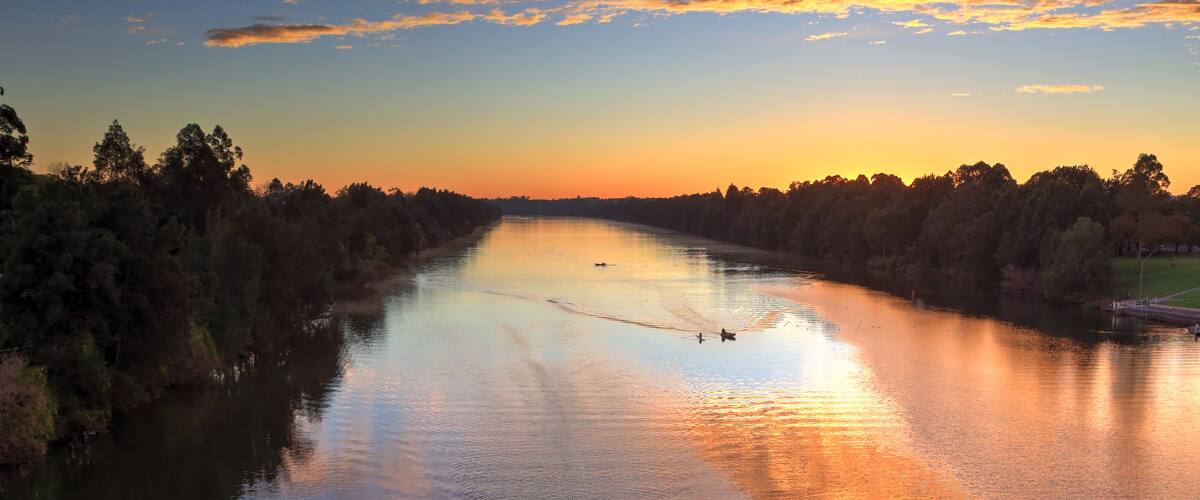 Un río al atardecer con un bote remando en primer plano y un puente al fondo.