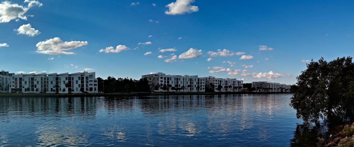 A river with buildings on the opposite bank, a clear sky with scattered clouds, and a bridge in the distance.