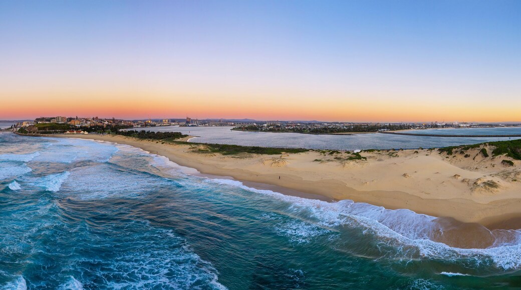 A coastal landscape with a sandy beach, waves, a lighthouse, and a distant city skyline at sunset.