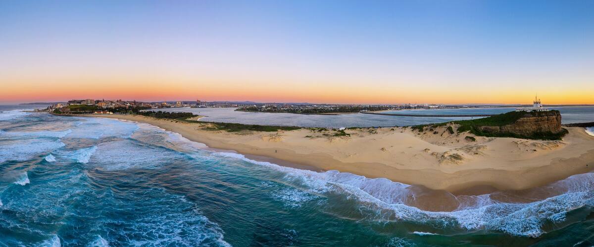 A coastal landscape with a sandy beach, waves, a lighthouse, and a distant city skyline at sunset.