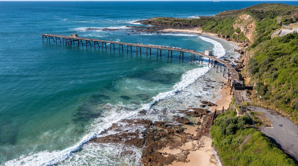A coastal landscape with a long pier extending into the sea, a sandy beach, rocky cliffs, and a clear sky.
