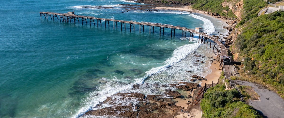A coastal landscape with a long pier extending into the sea, a sandy beach, rocky cliffs, and a clear sky.