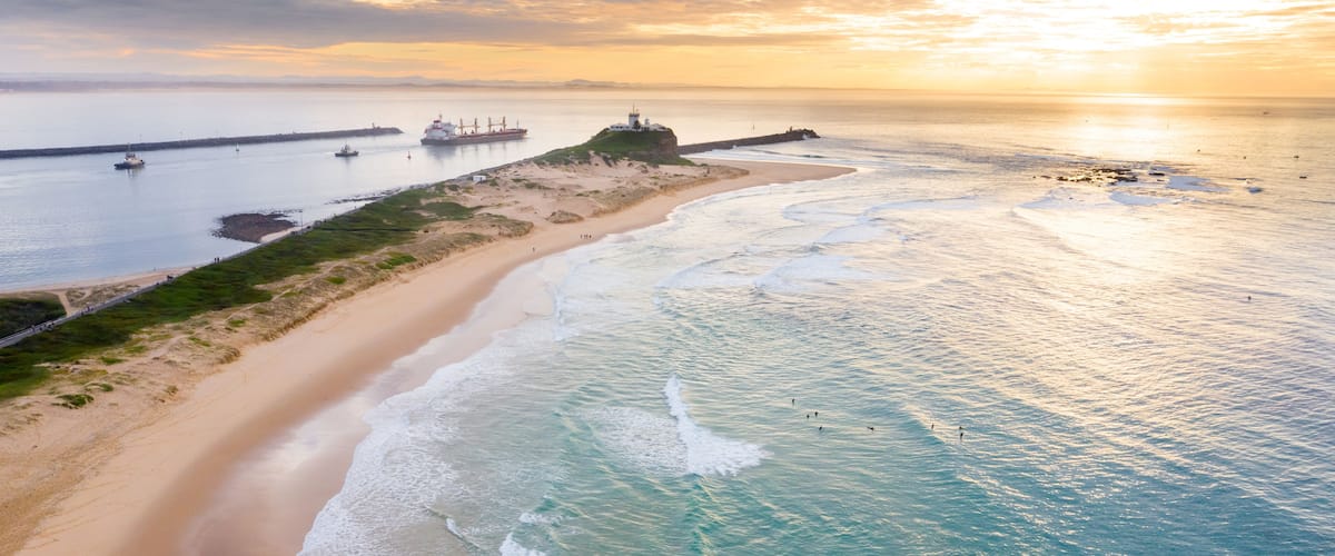 A coastal landscape with a sandy beach, ocean waves, a lighthouse, and a cargo ship in the distance.