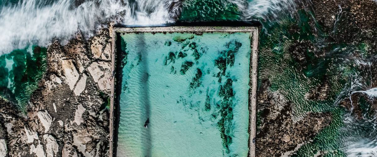 A turquoise swimming pool surrounded by rocky terrain and ocean waves.