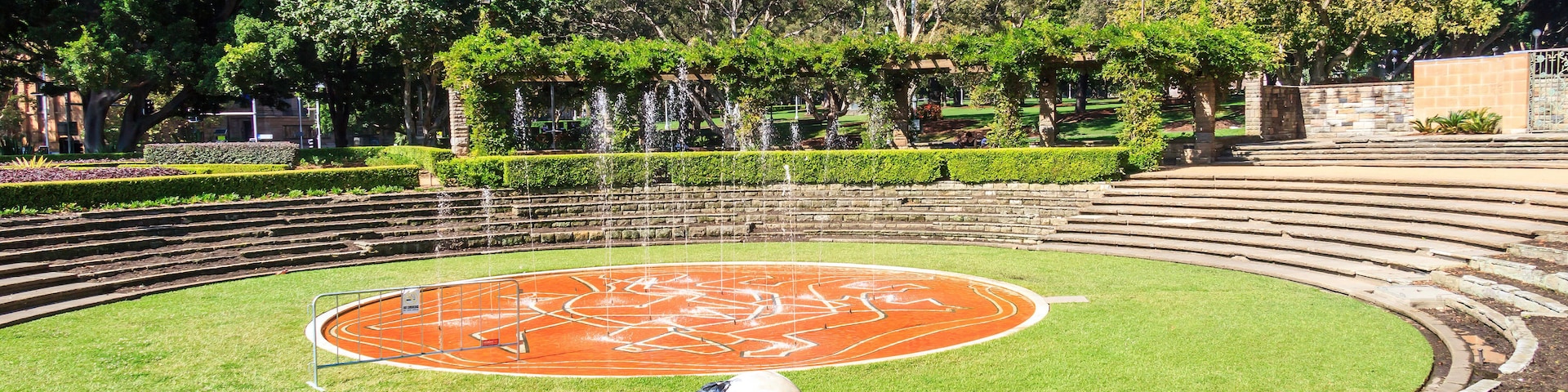 Ein Park mit einem runden Springbrunnen, grünen Hecken und Bäumen.