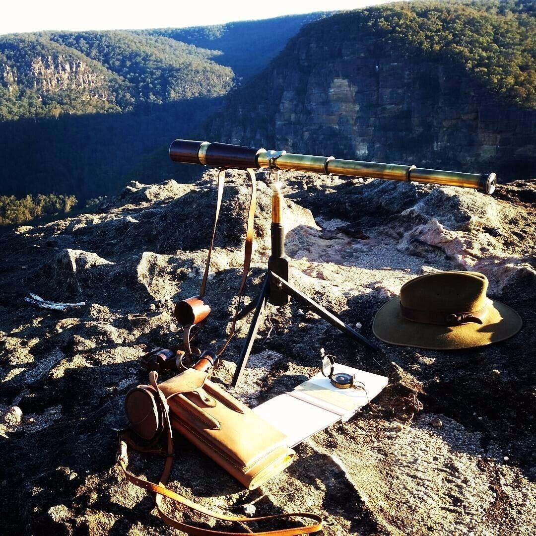 A telescope, a hat, a leather bag, and a notebook on a rocky surface with a mountainous backdrop.