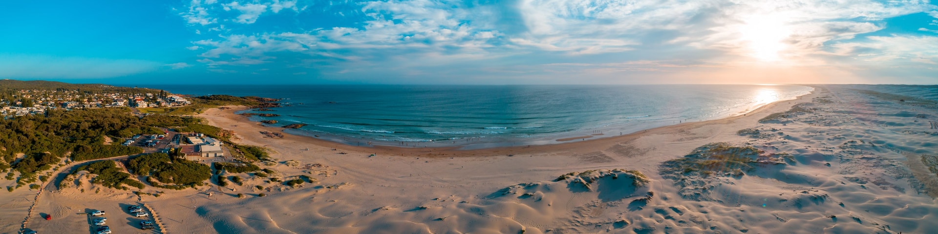 Paisagem costeira com uma praia arenosa, céu claro e uma cidade ao longe.