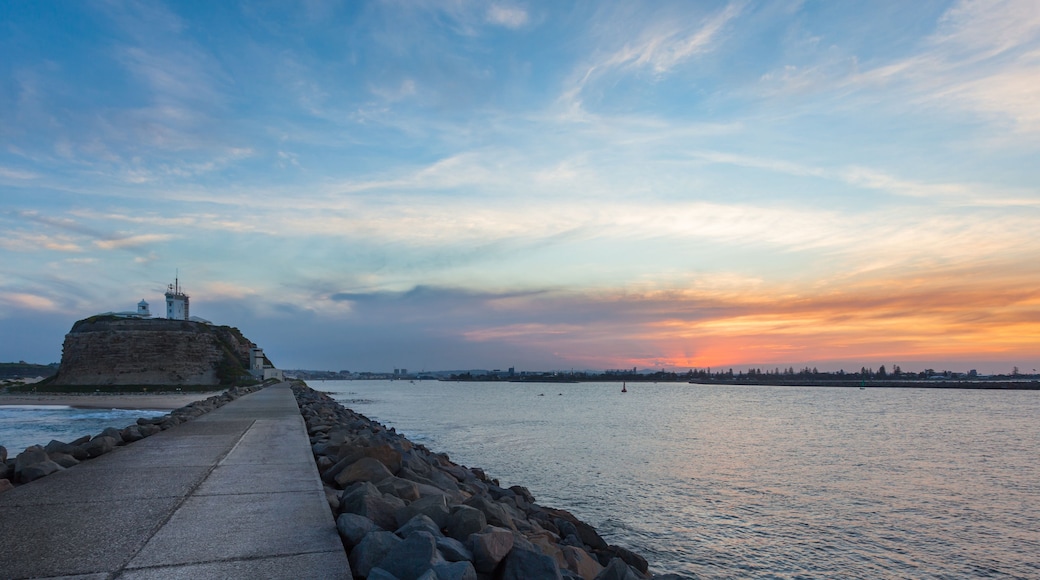 A coastal walkway leading to a lighthouse on a cliff, with a sunset in the background.