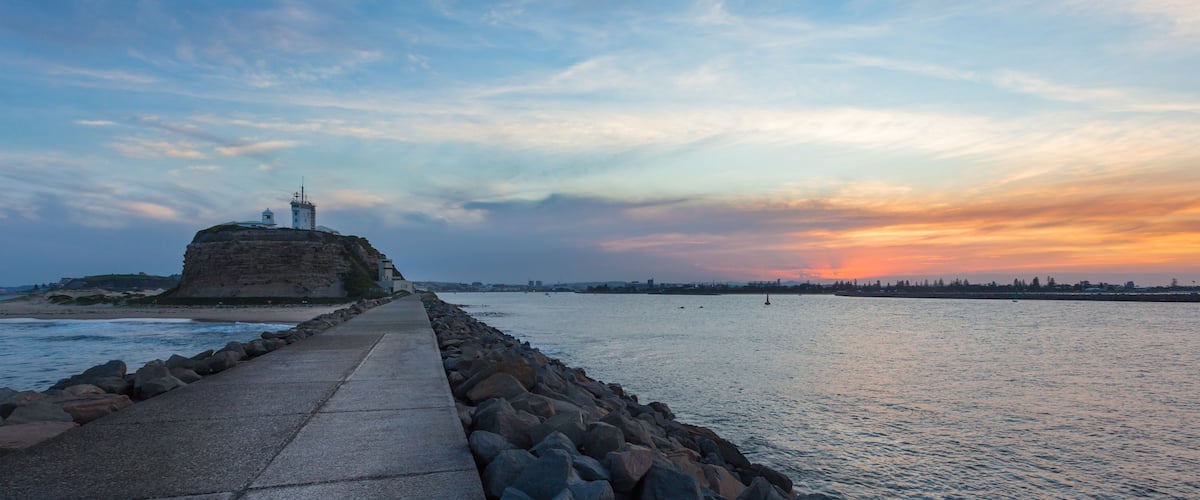 A coastal walkway leading to a lighthouse on a cliff, with a sunset in the background.