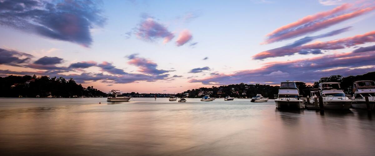 A calm harbor with boats at sunset, a clear sky, and a distant shoreline with buildings.