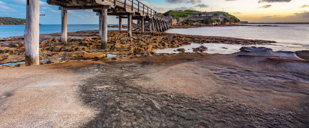 A coastal scene with a wooden pier extending into the sea, rocky shore, and a distant hill with buildings.