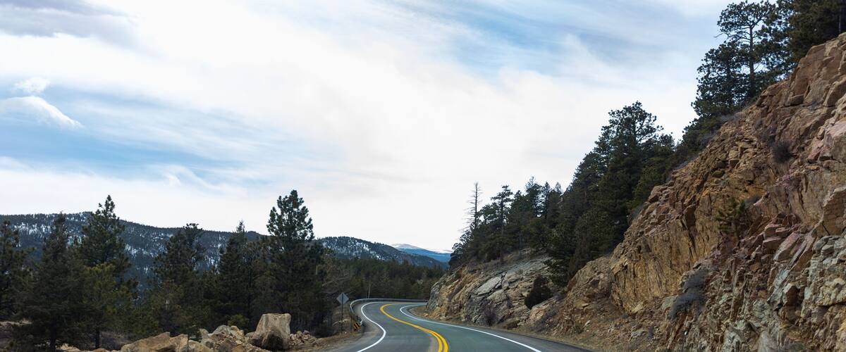 A winding mountain road with yellow center lines and white edge lines, surrounded by rocky cliffs and dense forests.
