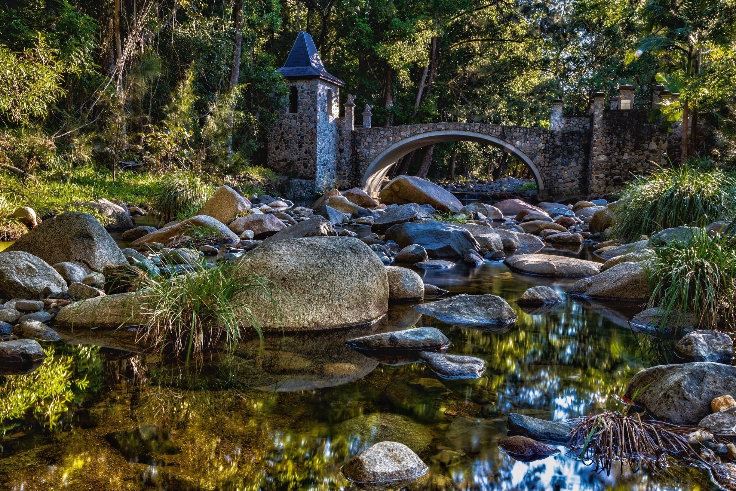 A stone bridge over a clear stream with large rocks and a small tower in the background.
