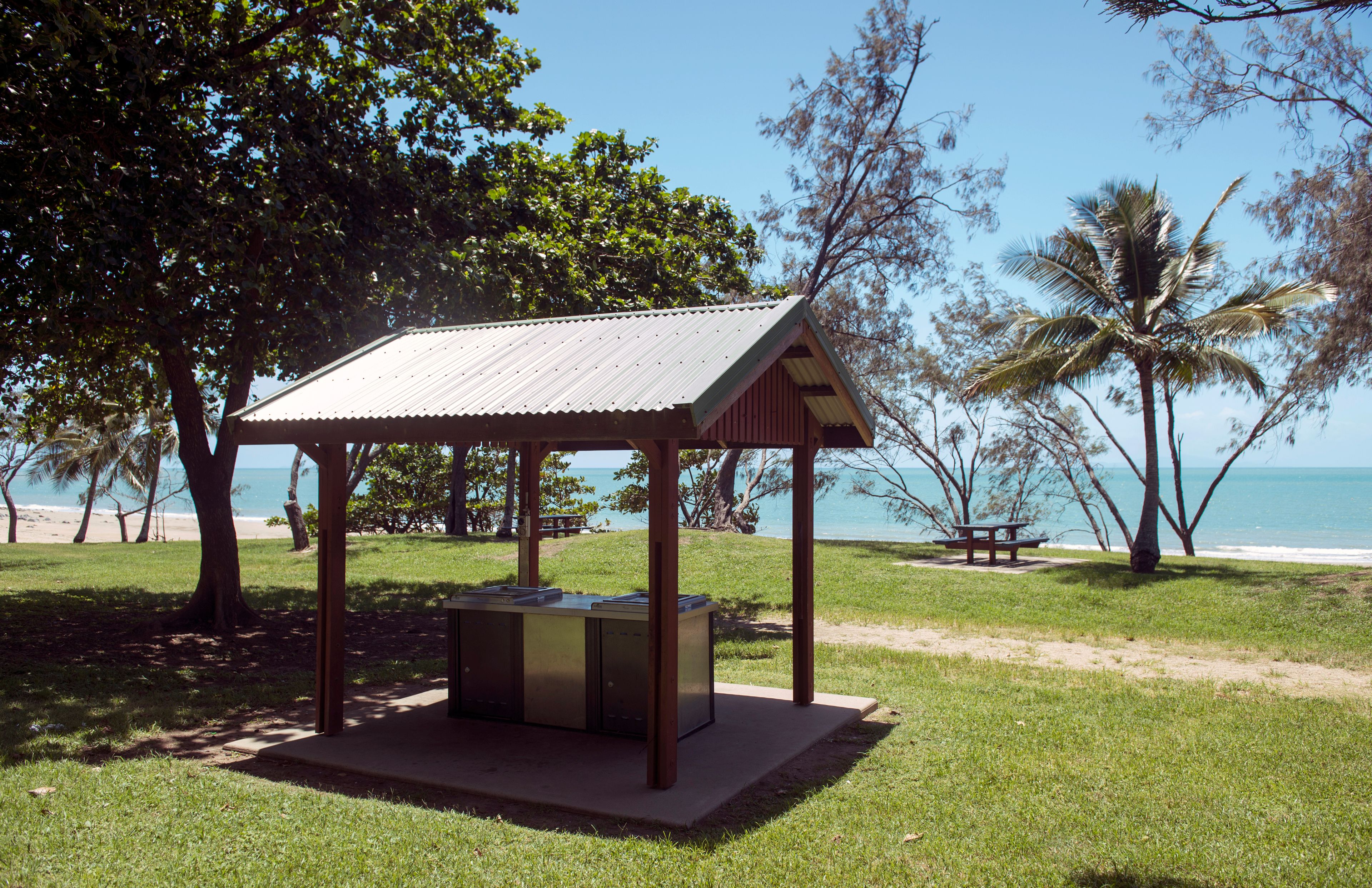A wooden gazebo with a metal roof, situated on a grassy area near a beach with palm trees and a clear blue sky.
