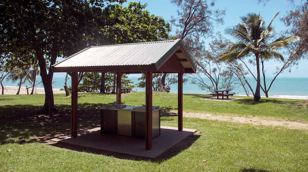 A wooden gazebo with a metal roof, situated on a grassy area near a beach with palm trees and a clear blue sky.