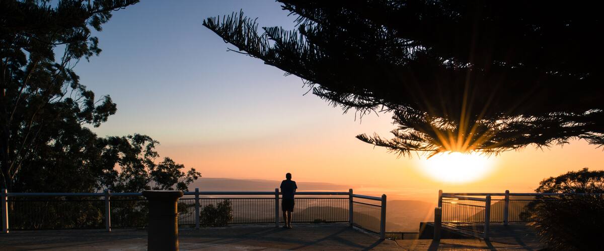 A sunset view from a park with a person standing on a viewing platform, overlooking a body of water.