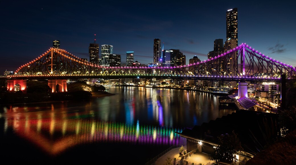 Un puente iluminado con luces de colores que se reflejan en el agua, un horizonte urbano con edificios iluminados y un cielo nocturno despejado.