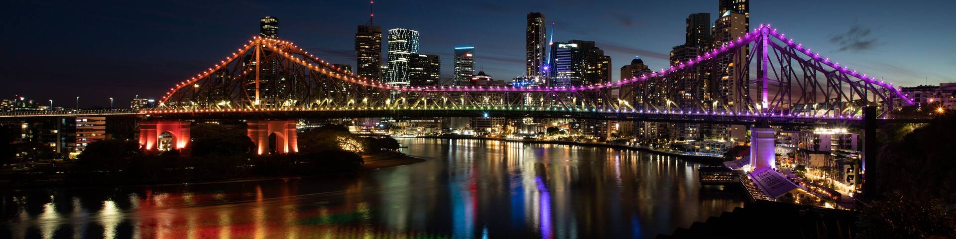 A brightly lit bridge with colorful lights reflecting on the water, a city skyline with illuminated buildings, and a clear night sky.