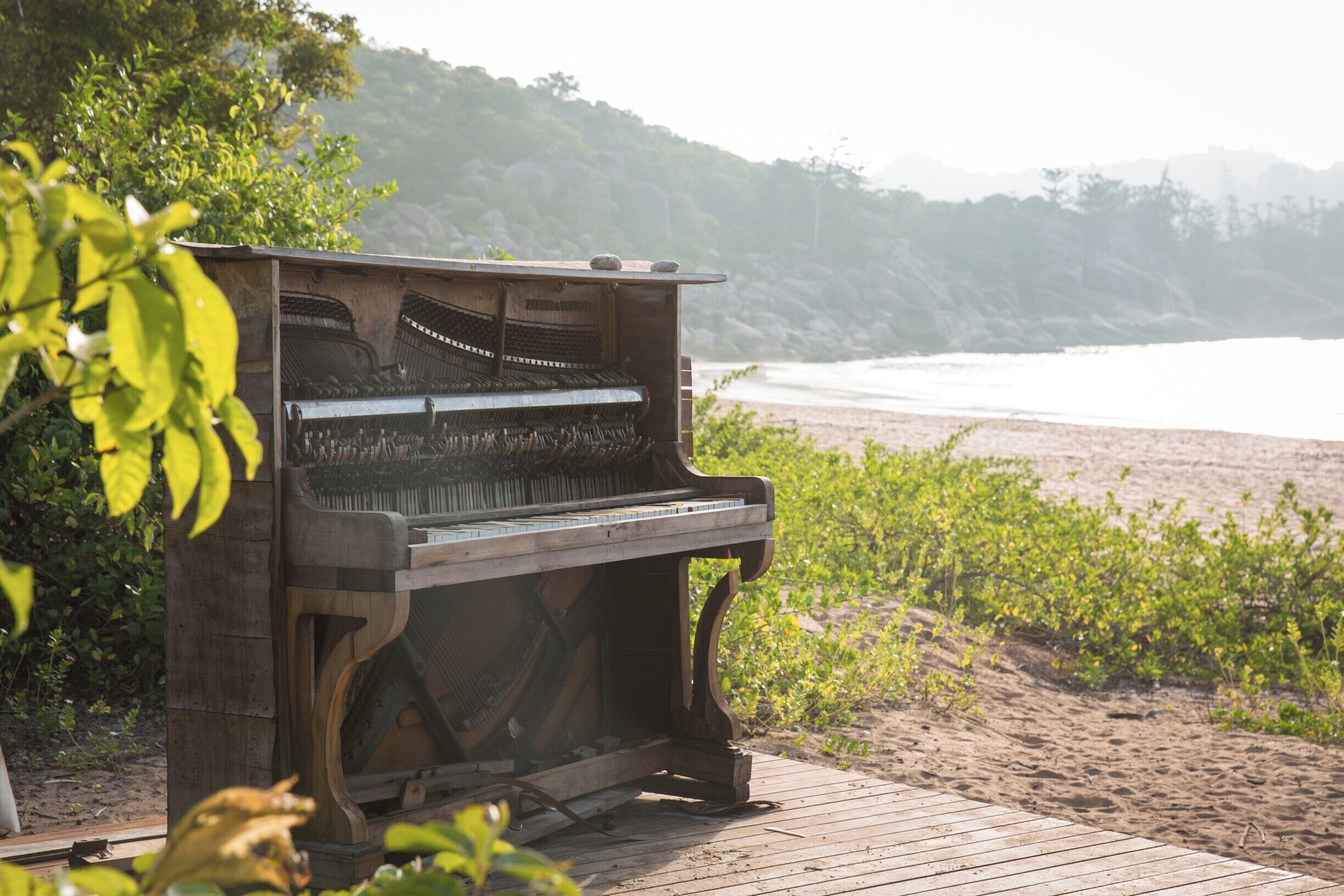 A grand piano on a wooden deck by the beach with a backdrop of hills and ocean.