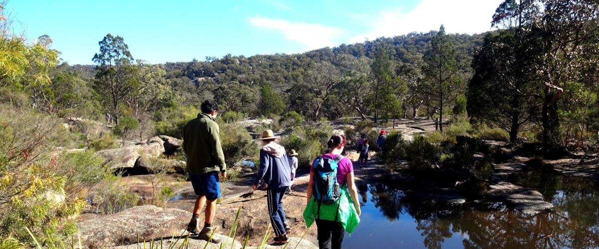 Hikers on a rocky path near a body of water with a forested hill in the background.