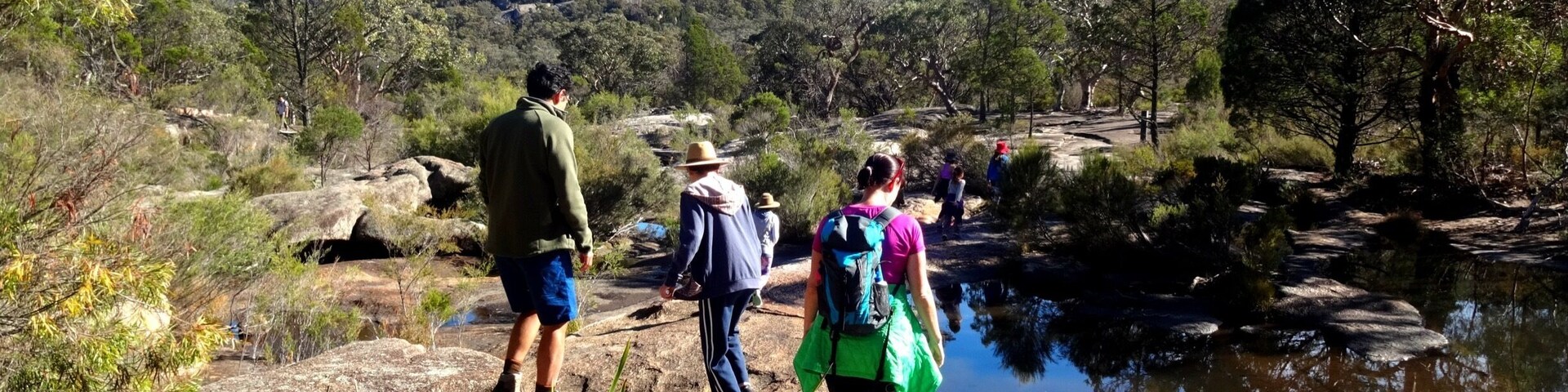 Hikers on a rocky path near a body of water with a forested hill in the background.