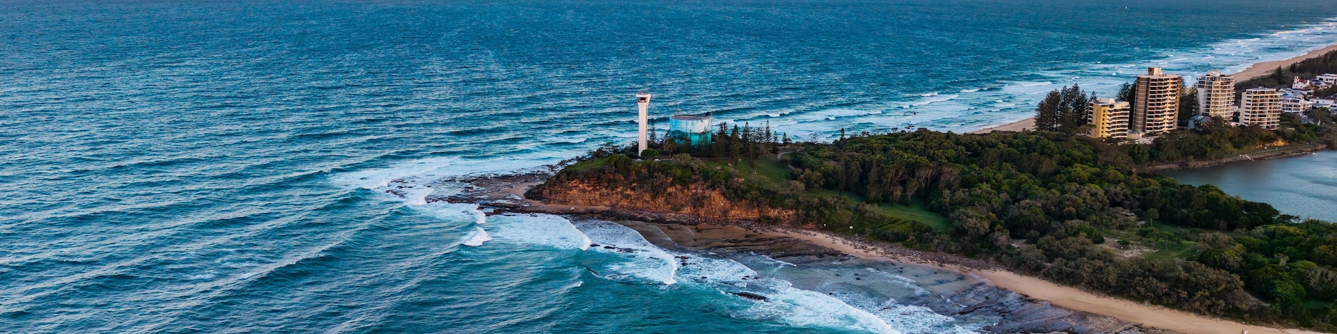 Paisagem costeira com um farol, praia e edifícios residenciais.