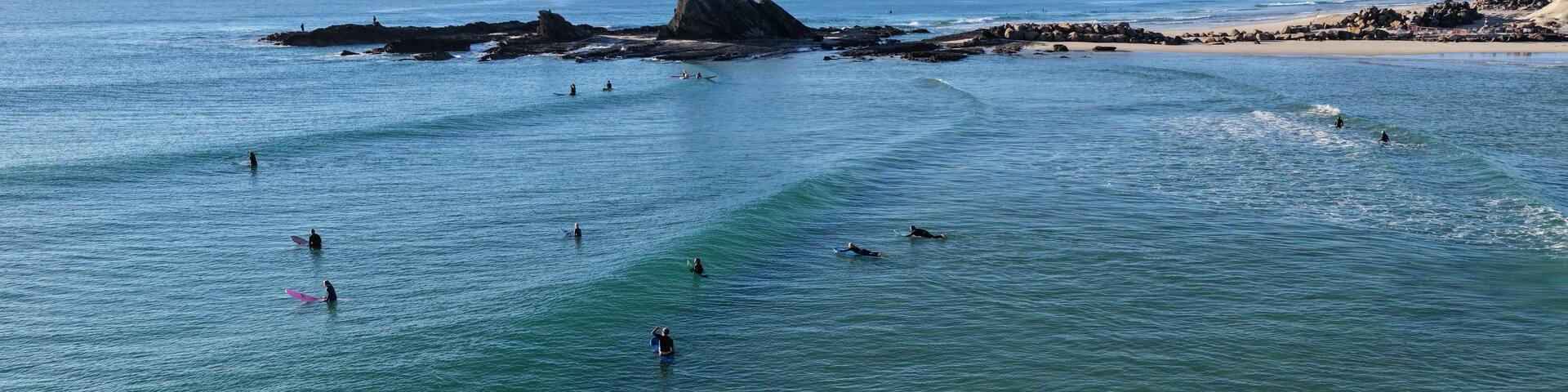 A coastal scene with surfers in the water, a rocky outcrop, and a distant city skyline.