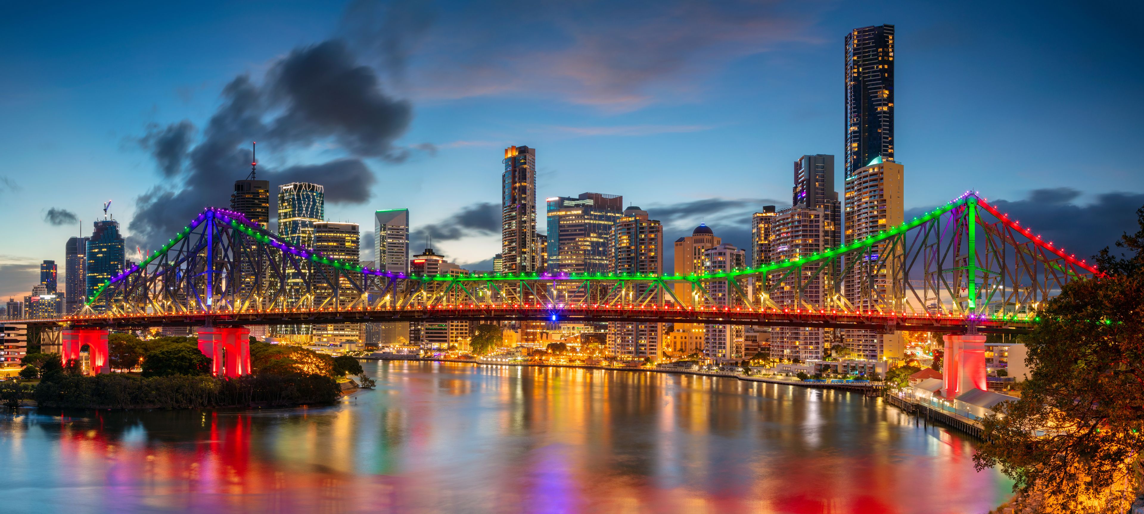 A brightly lit bridge with colorful lights spanning a river, surrounded by a city skyline with tall buildings.
