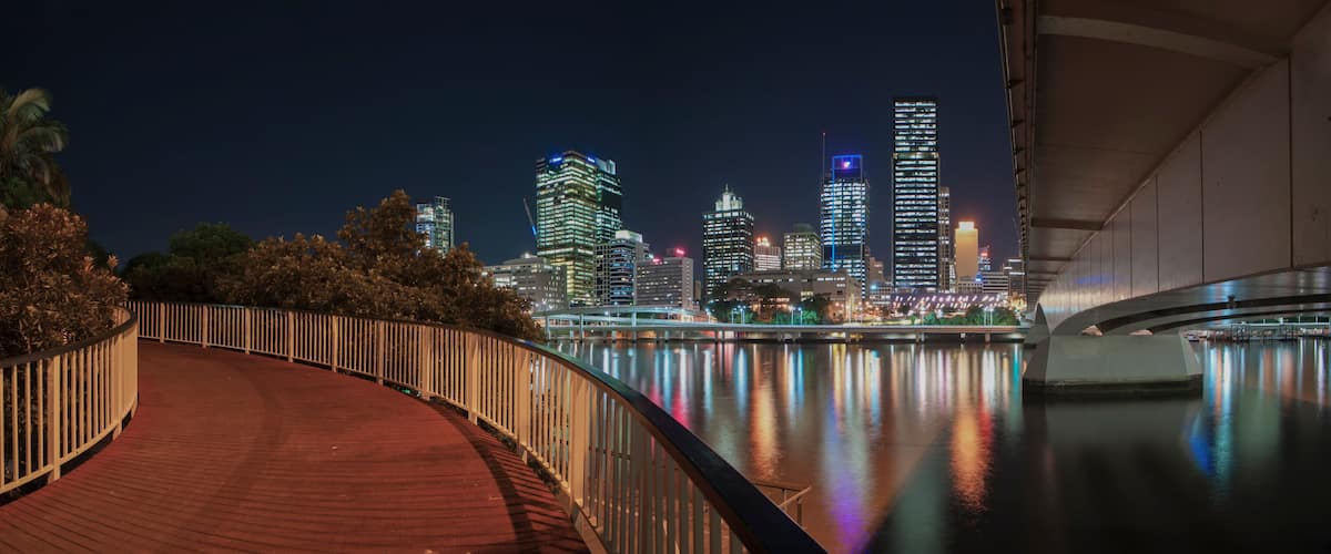 A nighttime cityscape with illuminated skyscrapers reflecting on a calm body of water, a curved wooden walkway, and a bridge in the foreground.