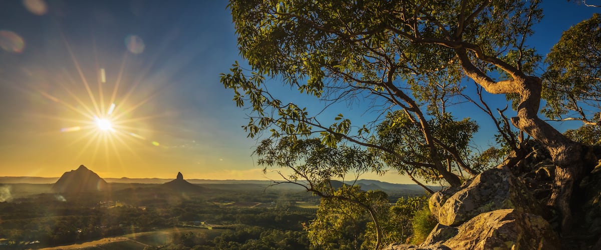 A mountainous landscape with a clear sky, a prominent sun, and a tree in the foreground.