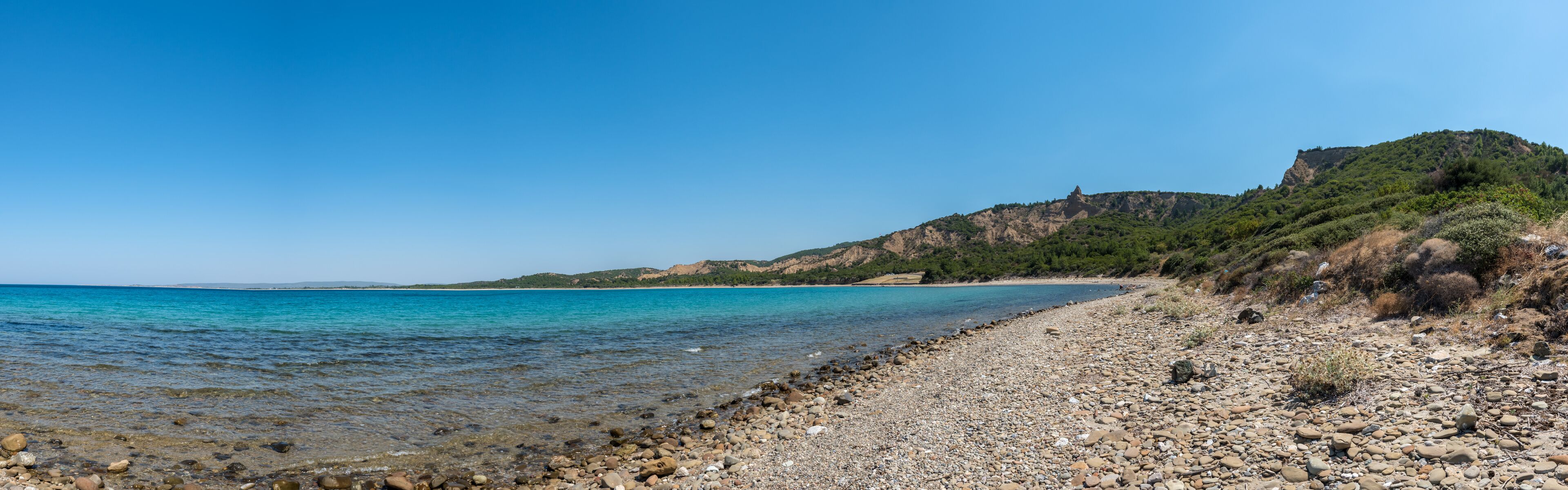 A pebble beach with clear turquoise water, a rocky shoreline, and a hill with sparse vegetation in the background.