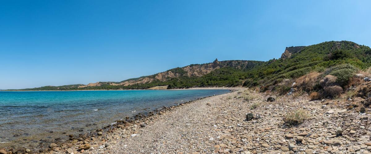 A pebble beach with clear turquoise water, a rocky shoreline, and a hill with sparse vegetation in the background.