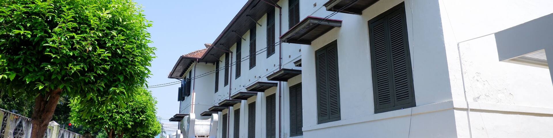 A row of white buildings with dark shutters, a tree-lined street, and a clear blue sky.