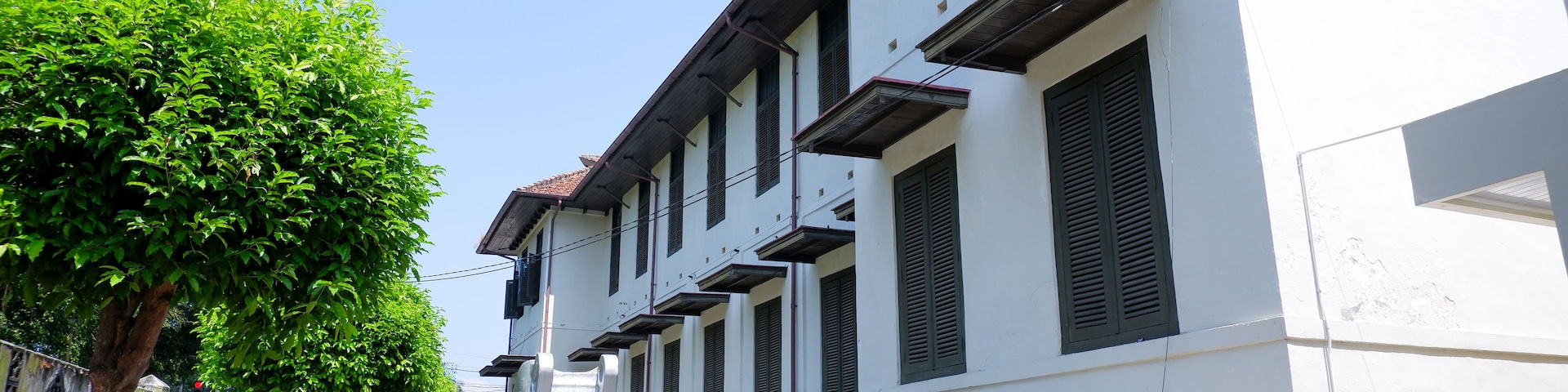A row of white buildings with dark shutters, a tree-lined street, and a clear blue sky.