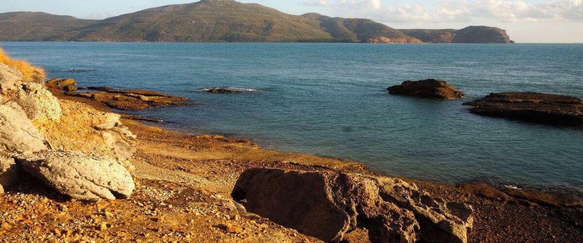 A coastal landscape with rocky shores, calm sea, and distant hills.