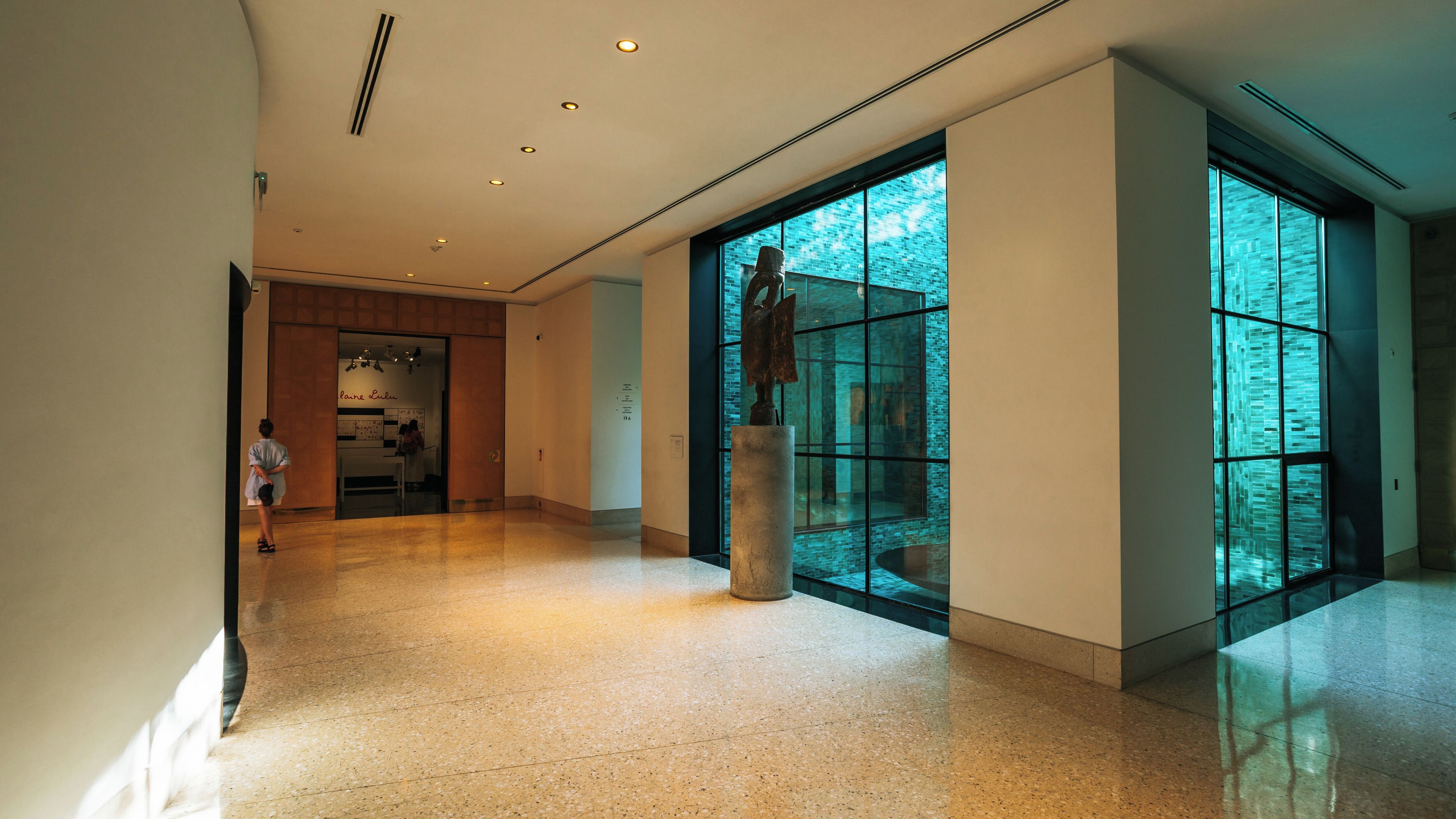 A spacious hallway with a polished floor, a large window with a view of a brick building, and a sculpture on a pedestal.